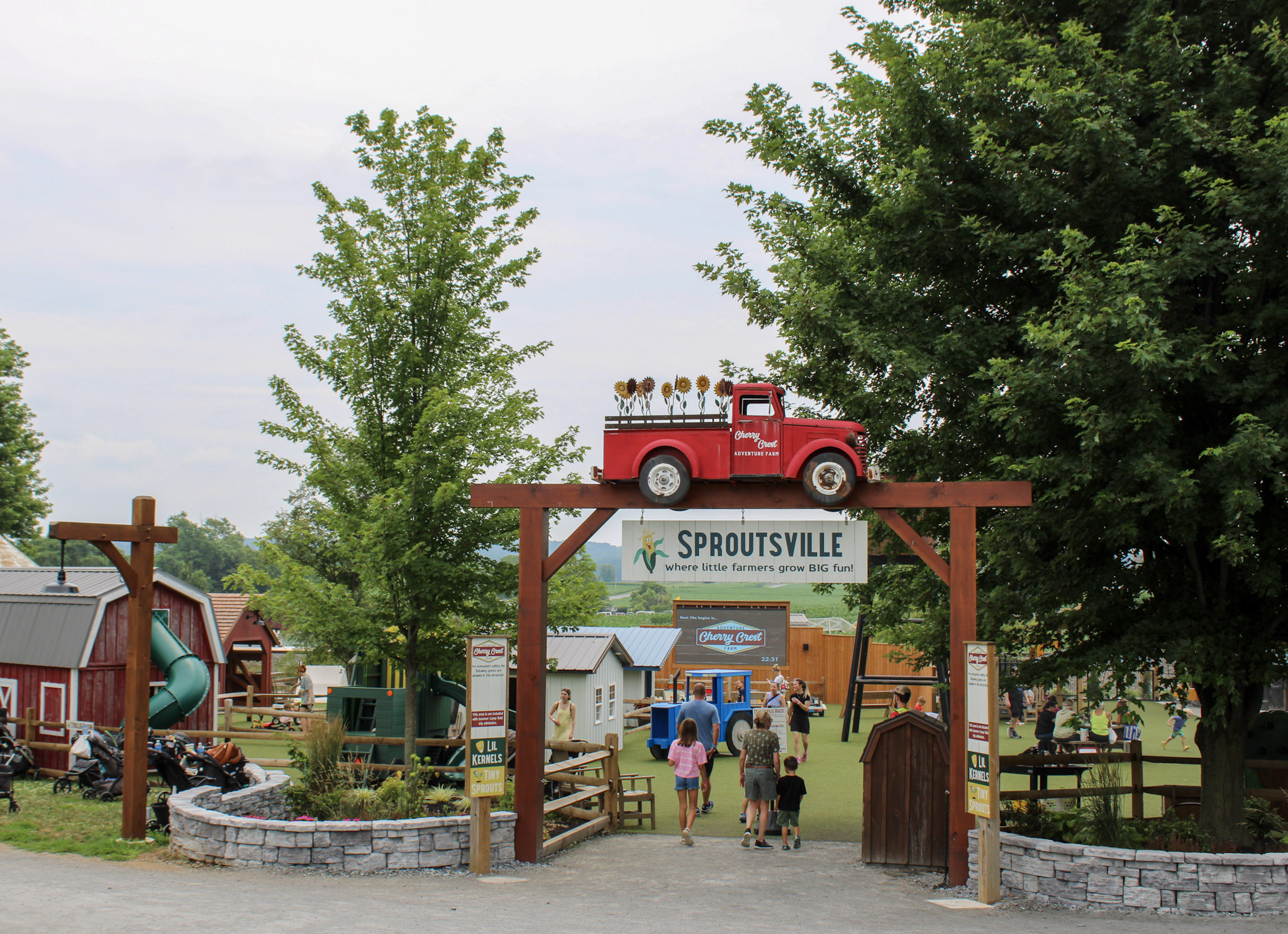 Sproutsville entrance sign with red pickup truck and farm buildings, children playing in the background.